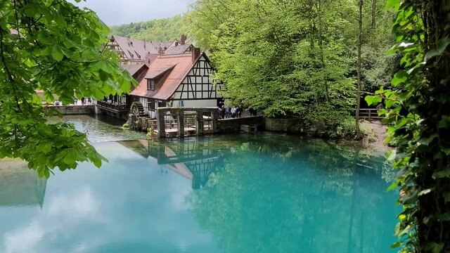 Scenic view of the natural river head called Blautopf in Blaubeuren, Germany. A historic hammer mill reflects in the blue-turquoise water.
