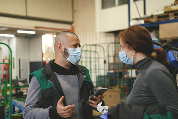 Industrial workers with face masks protected against corona virus discussing about production in factory. People working during COVID-19 pandemic.