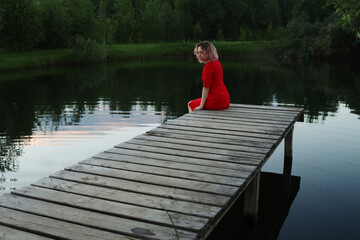 Fototapeta na wymiar Young attractive woman in a red tight dress sits on the bridge by the pond at sunset. Rustic romance. Life in the countryside