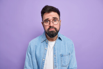 Handsome frustrated bearded man with pity expression frows face looks unhappily being bothered faces troubles wears round spectacles denim shirt isolated over purple background. Negative emotions
