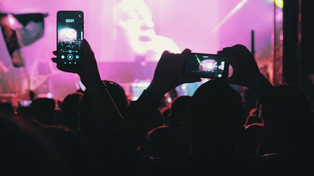 People filming rock concert on smartphones. Silhouettes crowd of people dancing at an open-air music festival. Fans applaud, raise hands up, and photographing. Party People in Action. 4K, Slow Motion