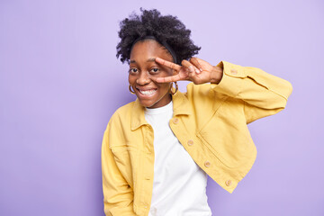 Horizontal shot of cheerful carefree dark skinned woman makes peace gesture over eye smiles broadly as fun shows disco sign stands well dressed against purple background. Body language concept