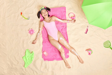 Obraz na płótnie Canvas Carefree smiling Asian teenage girl with perfect figure wears sunbathing suit poses on soft pink towel applies sunscreen on body while sunbathing spends leisure time at tropical sandy coastline