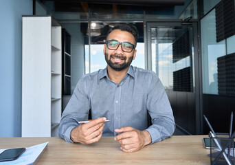 Smiling indian businessman in glasses talking to camera at work by video call conference. Financial advisor executive consulting client remotely online in modern office looking at camera. Webcam view