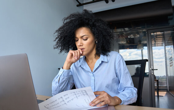 Concerned African American businesswoman ceo analyst sitting at desk reading papers financial reports with laptop in contemporary corporation office. Financial data analyst working with big data.