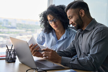 Multiethnic male indian mentor and female African American intern sitting at desk with laptop doing paperwork together discussing project financial report. Corporate business collaboration concept. - obrazy, fototapety, plakaty