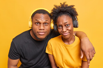 Fototapeta premium Photo of friendly dark skinned woman and man embrace and look attentively at camera dressed casually listen music via headphones isolated over vivid yellow background learn foreign languages together