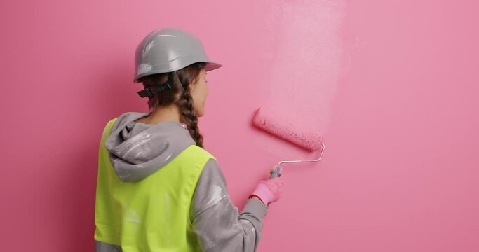 Back view of happy female painter dances while applying paint on wall uses painting roller does home improvement wears helmet reflective vest paints or refurbishes new apartment. DIY concept