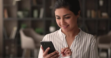 Attractive Indian woman standing alone indoor holding in hands smart phone dial number on touchscreen. Cheerful young female surfing internet, websurfing website, choose buying goods on-line concept - Powered by Adobe