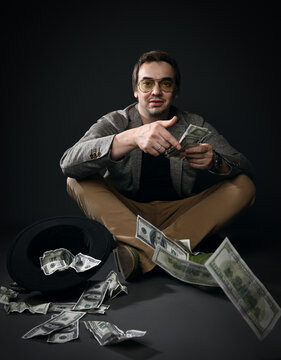 Rich insolent middle-aged man in jacket, jeans and shoes sits on floor by upside down hat full of dollars throwing cash, spending over dark gray background