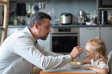 Fototapeta premium A caring man with a beard feeds his little daughter. A little girl sits at a children's dining table and dines with her dad. Happy fatherhood.