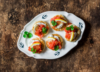 Delicious tapas, appetizer-mini pancakes with salmon, cream cheese and lemon on a wooden background, top view
