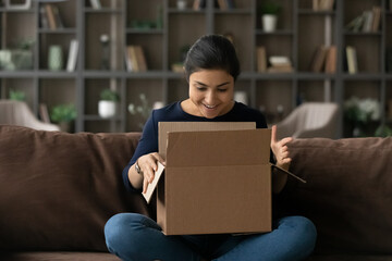 What a nice surprise. Joyful teenage indian female addressee sit on sofa looking inside big cardboard box received by mail service. Happy interested young woman opening carton postal parcel enjoy gift - obrazy, fototapety, plakaty