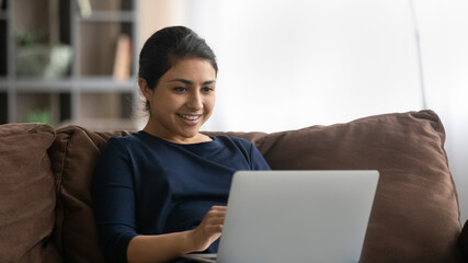 Happy young indian lady enjoy weekend rest on sofa chat by video call using wifi internet on laptop watch good comedy movie. Joyful teen female relax at home read funny blog article on computer screen