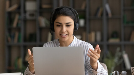 Confident millennial indian female coach tutor in modern headphone set explain academic program to remote students online. Young woman teacher speak give virtual lesson lecture before laptop webcam