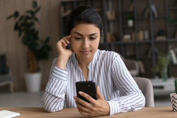 Serious millennial mixed race woman sit at desk read text on smartphone screen use mail client to...