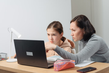 A happy family. Girl and mother are doing homework while sitting at a laptop in the room. Mom teaches daughter sitting at the table at home.