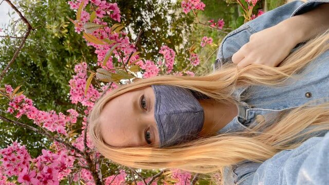 Vertical video. Voman wearing a facemask makes selfie video standing under blooming sakura tree. Travel to Japan in a times of pandemics. Shot on a phone