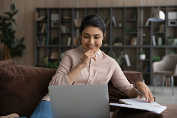 Web education. Indian teen female study from home on cozy sofa using laptop portable headphones....