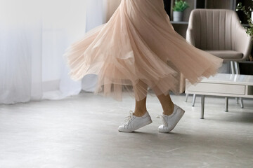 Young lady in light skirt white sporty shoes on feet dance move on tiptoes at light spacious living room. Close up of teen female dancer slender legs expressing positive emotions in motion. Copy space