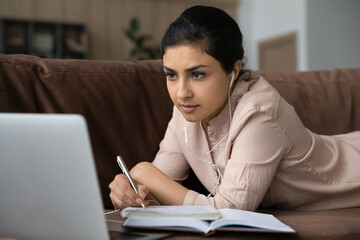 Focused indian female student in wired headphones lie on sofa in comfy pose before laptop screen...