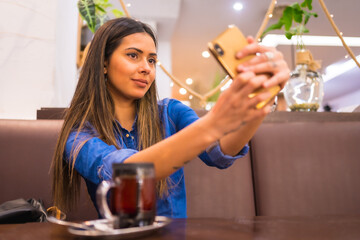 Street Style in the city, a brunette Caucasian girl in a denim shirt having tea in a coffee shop. Taking a selfie with the phone
