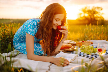Fototapeta premium Beautiful woman sitting on the blanket reading a book and smiling outdoors. People, lifestyle, relaxation and vacations concept. Picnic set with fruit, cheese, baguette.