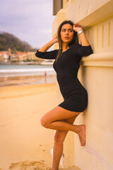 Lifestyle, pretty young caucasian brunette with a short black dress, summer vacation, looking at camera with sexy look and enjoying the beach