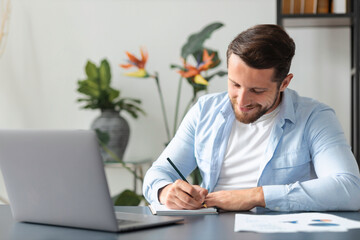 Concentrated caucasian man taking notes while sitting at table with laptop at home or office, young...