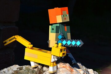Naklejka premium LEGO Minecraft large action figure of Alex with diamond sword standing on rock with friendly Ocelot cat, dark background. 
