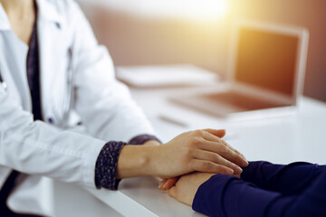 Unknown woman-doctor is holding her patient's hands, discussing current health examination, while they are sitting at the desk in the sunny cabinet in a clinic. Physician with a stethscope at work