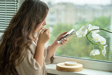 Girl drinking coffee looking at rain through a window at home or hotel and holding phone. Girl with...
