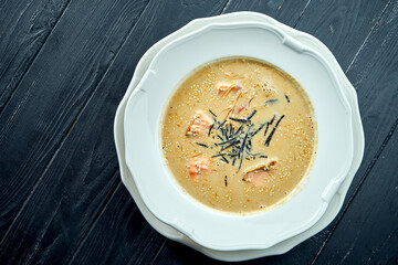 Appetizing coconut soup with salmon, nori and sesame seeds, served in a white plate on a dark wood background. Pan-Asian cuisine