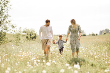 Fototapeta premium Cheerful parents having fun with their little son 3 years in summer field while holding hands and walking together.