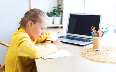 Caucasian schoolgirl in a yellow sweatshirt is doing homework at home in front of a laptop, writes in a notebook. Home learning, e-learning. Back to school. Cozy workplace by the window.