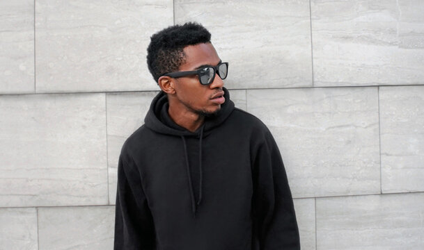 Portrait close up of stylish young african man posing looking away wearing a black hoodie, sunglasses on a city street over gray wall background