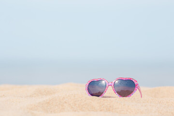 Fototapeta na wymiar Baby girl sunglasses with pink hearts frame on sea beach sand in hot sunny summer day. Closeup. Front view. Empty place for positive text, inspirational quote or sayings on blue sky background.