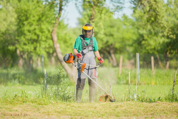 One young adult man in protective clothes mowing green grass with gasoline lawn trimmer at garden...