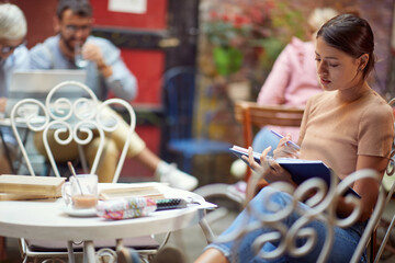 young adult female sitting in outdoor cafe reading a book alone, with other people in the background.