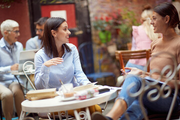 two caucasian young adult women sitting in the outdoor cafe, talking, socializing, smiling, looking each other