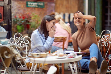young adult caucasian woman telling shyly gossip to her female friend sitting in the outdoor cafe