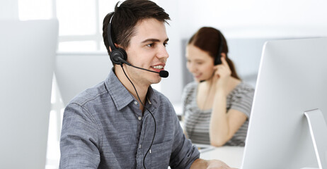 Casual dressed young man using headset and computer while talking with customers online. Group of...
