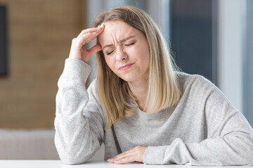 Portrait of a young blonde woman sitting at a table in the office or at home and holding his head because of severe pain. Headache or migraine or premenstrual fever indoors. In casual clothes
