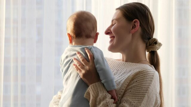 Happy smiling mother hugging and kissing her little baby son at big window in living room. Concept of family happiness and parenting