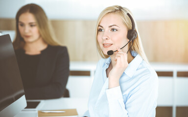 Blonde business woman sitting and communicated by headset in sunny call center office