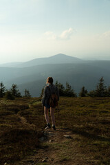 Fototapeta premium Woman on the summit of Králický Sněžník between Poland and the Czech Republic, at sunset, high mountain meadow, with Czarna Gora in the background. Classic hiking with a backpack.