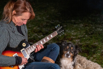 Young woman plays guitar in the campsite by the campfire. In casual wear, wool vest, jeans and outdoor boots. Her Australian Shepherd is watching and listening to the music
