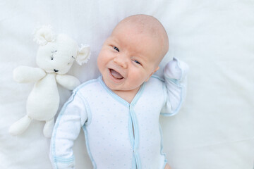 smiling or laughing newborn baby boy lying on the back of the crib on the cotton bed at home before going to bed with a soft toy, baby week, the concept of birth and infancy, top view