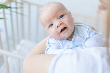 a newborn baby boy in his mother's arms smiles near the crib at home, parental love and care for the baby in the first days after birth