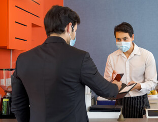 The cashier in the apron and face mask hands a receipt to a caucasian customer. A young businessman...